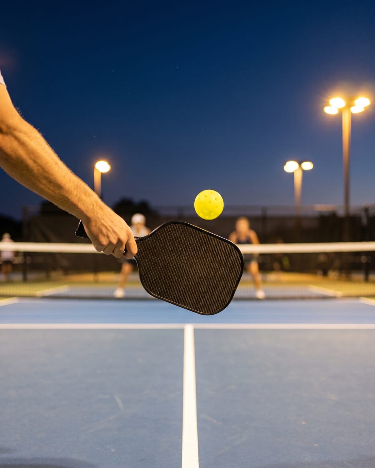 Carbon-fiber pickleball paddle meeting a yellow ball at the kitchen line on a floodlit court at dusk